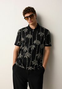 Polo negru cu imprimeu alb de palmieri, mâneci scurte și guler cu fermoar. Modelul poartă ochelari de soare negri și pantaloni închiși la culoare.