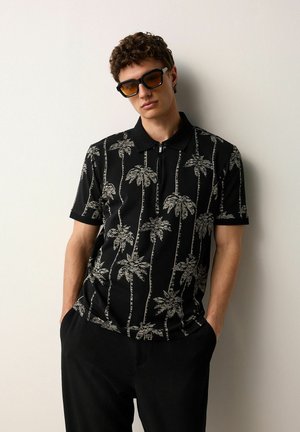 Camisa polo negra con estampado de palmeras blancas, mangas cortas y cuello con cremallera. El modelo lleva gafas de sol negras y pantalones oscuros.