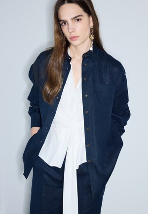 Joven mujer con cabello largo y castaño que lleva pendientes dorados de gota, una camisa azul marino abotonada sobre una blusa blanca atada y pantalones azul marino a juego.
