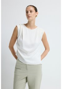 Hvid ærmeløs top i blødt, flydende stof med subtile plisseringer; parret med lysegrønne skræddersyede bukser.