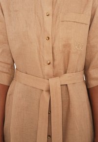 Linnen skjortekjole i beige med korte ermer, frontknapper, en brystlomme og belte i livet; har broderte Initialer "MM."