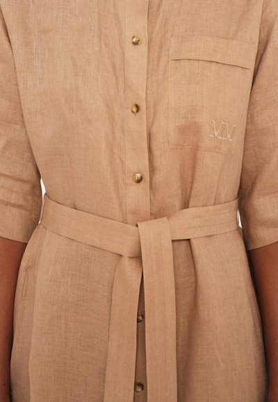Vestido camisola em linho bege, com mangas curtas, botões frontais, um bolso no peito e cintura com cinto; apresenta iniciais bordadas "MM."