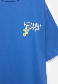 T-shirt en coton bleu à manches courtes, avec un imprimé graphique blanc et coloré de "Nectar Hills Peachys" et des icônes de fruits ludiques sur la poitrine.