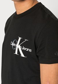 Černé bavlněné tričko s kulatým výstřihem, na přední straně se nachází velké bílé logo "Calvin Klein Jeans" a stylizovaný design "CK".