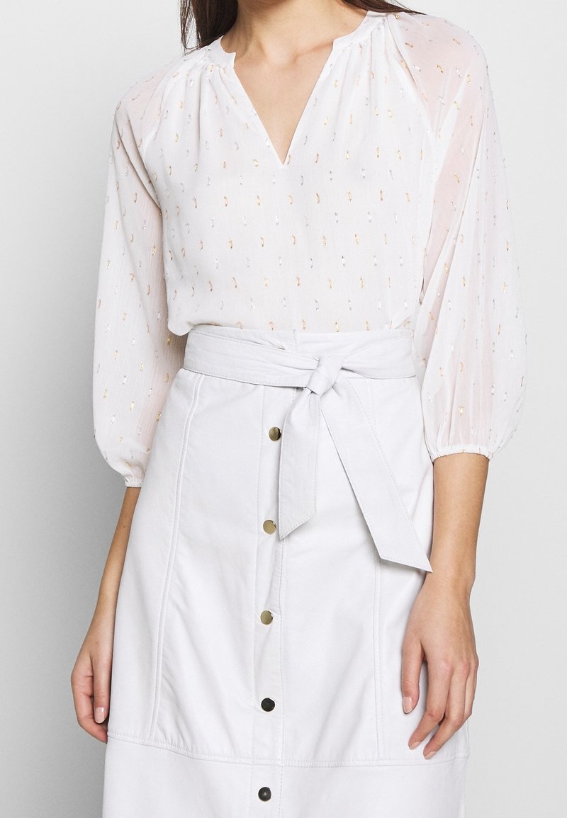 Blusa bianca semi-trasparente con scollo a V e maniche a palloncino, con delicati accenti metallici. Abbinata a una gonna bianca a vita alta.