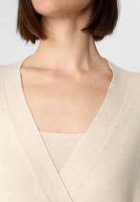 Maglione leggero beige con scollatura a V, caratterizzato da una sottile trama e indossato sotto un top abbinato. Tessuto morbido e liscio.