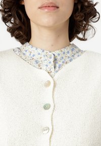 Světlý krémový kardigan s pěti knoflíky, pod ním květinově vzorovaná košile s límečkem, která ukazuje jemné textury a kontrastní designy.