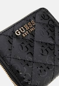 Portefeuille noir matelassé avec une finition brillante et texturée. Présente un logo doré "GUESS EST 1981" et une fermeture éclair. Design lisse et élégant.