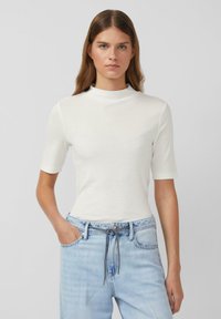 Biała krótka bluzka z mock neck, obszyta prążkowanym materiałem, dopasowany krój, zestawiona z jasnoniebieskimi dżinsami z elastycznym paskiem.