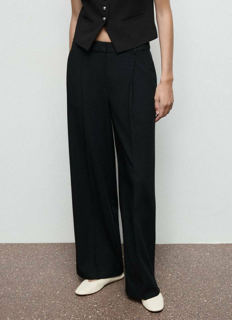 Zwarte wijde broek met plooien, gemaakt van een gladde stof. Gecombineerd met een aansluitend zwart vest met knoopdetails.