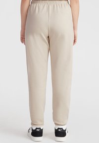 Beige joggers met elastische taille en taps toelopende pijpen, gemaakt van zacht materiaal, gecombineerd met zwart-witte sportschoenen. Geen zichtbare zakken.