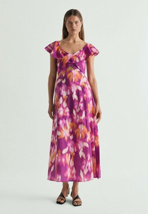 Mujer de pie con un vestido maxi floral en tonos morado, naranja y blanco, con mangas acampanadas, escote en V y sandalias negras sobre un fondo liso.