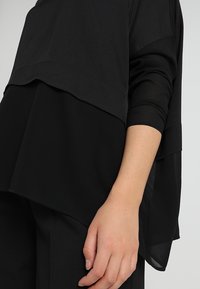 Top nero a strati realizzato in un tessuto leggero e texture, con maniche lunghe e un design morbido; presenta un orlo con lunghezze variabili.