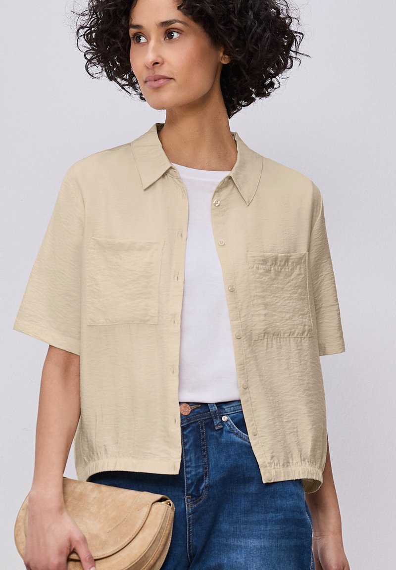 Femme aux cheveux bouclés portant une chemise beige à manches courtes et boutonnée par-dessus un haut blanc et un jean bleu, tenant une pochette en daim beige.