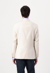 Beige blazer in lightweight fabric features a straight back design, single button closure, and three-button cuffs. Worn over a light blue shirt.