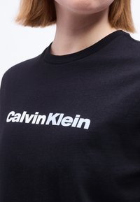 Κοντινή λήψη ενός ατόμου που φοράει μαύρο T-shirt με το λογότυπο "Calvin Klein" σε λευκό κείμενο στο στήθος.