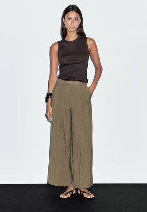 Braunes ärmelloses Oberteil kombiniert mit weiten olivgrünen Hosen. Das Modell steht in schwarzen Sandalen mit einem klobigen schwarzen Armband an ihrem Handgelenk.