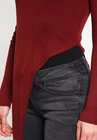 Bordeauxfarbiges, geripptes Langarmoberteil mit asymmetrischem Saum, kombiniert mit schwarzen Skinny-Jeans, die ein Design mit Fronttaschen und leichte Ausbleichungen aufweisen.