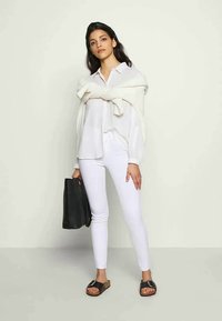 Camisa branca de mangas longas, com botões, sobre uma blusa branca, combinada com jeans skinny brancos e sandálias de slide castanhas. Bolsa preta na mão.