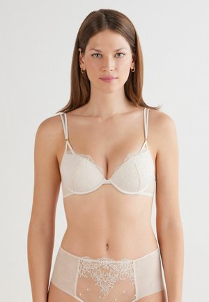 Soutien-gorge en dentelle beige avec bretelles ajustables, présentant des motifs floraux et un rembourrage doux. Culottes hautes assorties en tissu transparent avec des détails brodés.