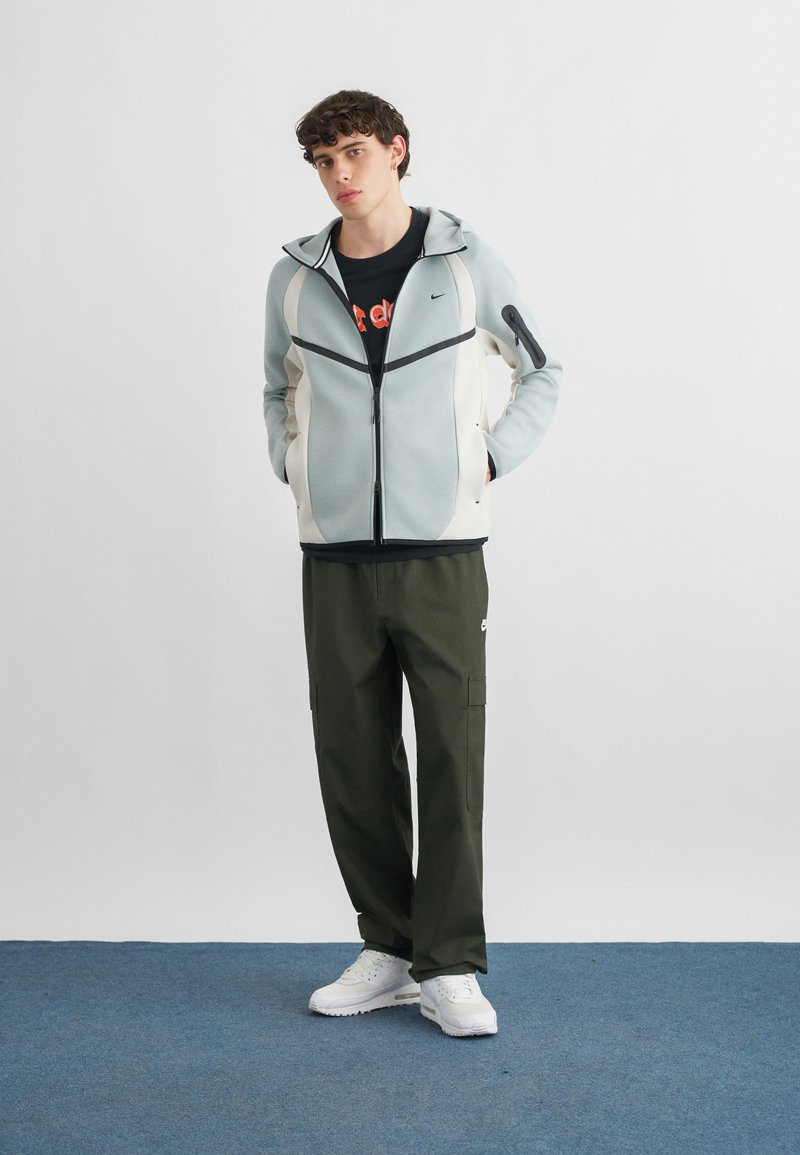 Junger Mann steht mit den Händen in den Taschen, trägt einen hellgrauen Kapuzenpullover mit Reißverschluss, ein schwarzes Grafik-T-Shirt, olivfarbene Cargohosen und weiße Sneakers auf blauem Teppich.