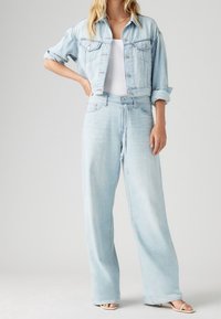 Mujer vestida con una chaqueta de mezclilla corta de color azul claro con las mangas remangadas, jeans de pierna ancha de cintura alta, una camiseta sin mangas blanca y sandalias de tacón beige.