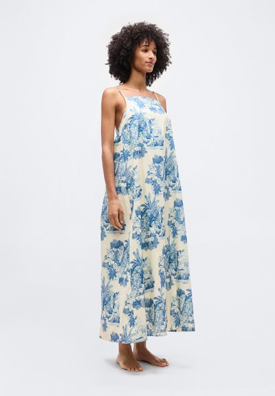 Γυναίκα με σγουρά μαλλιά, όρθια, ξυπόλυτη, που φοράει ένα αμάνικο, μακρύ μέχρι το πάτωμα λευκό φόρεμα με μπλε λουλουδάτα και τοπία μοτίβα.