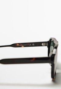 Svarta sköldpaddsmönstrade solglasögon med tjocka bågar, gröna linser och ingraverade detaljer på skalmarna som indikerar UV-skydd och certifiering.
