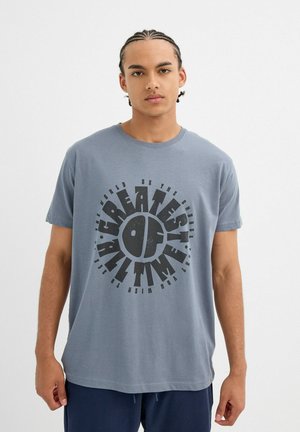 Camiseta de algodón gris con un audaz diseño gráfico negro con las palabras "EL MEJOR DE TODOS LOS TIEMPOS" en un diseño circular. Mangas cortas y cuello redondo.