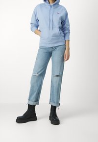 Un sweat à capuche bleu clair avec une poche avant et un cordon de serrage ajustable. Porté avec un jean bleu clair déchiré et des bottes de combat noires à semelles épaisses.