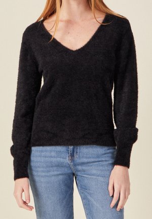 Femme portant un pull noir duveteux à col en V et manches longues, avec un jean bleu clair, montrée des épaules aux hanches, debout devant un fond uni.