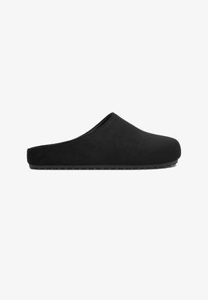 Zapatilla negra de tipo slip-on con puntera cerrada y redondeada, talón bajo y suela de goma texturizada para uso en interiores.