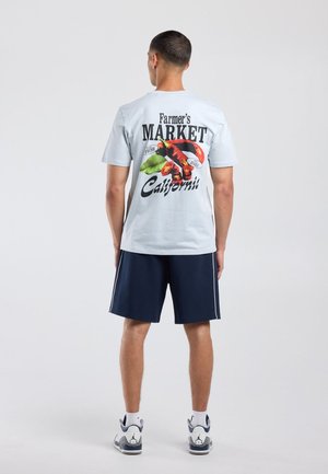 Man staat met zijn rug naar voren gekeerd, draagt een lichtblauw T-shirt met "Farmer's Market California" en chilipeperafbeeldingen, marineblauwe shorts en witte sneakers.