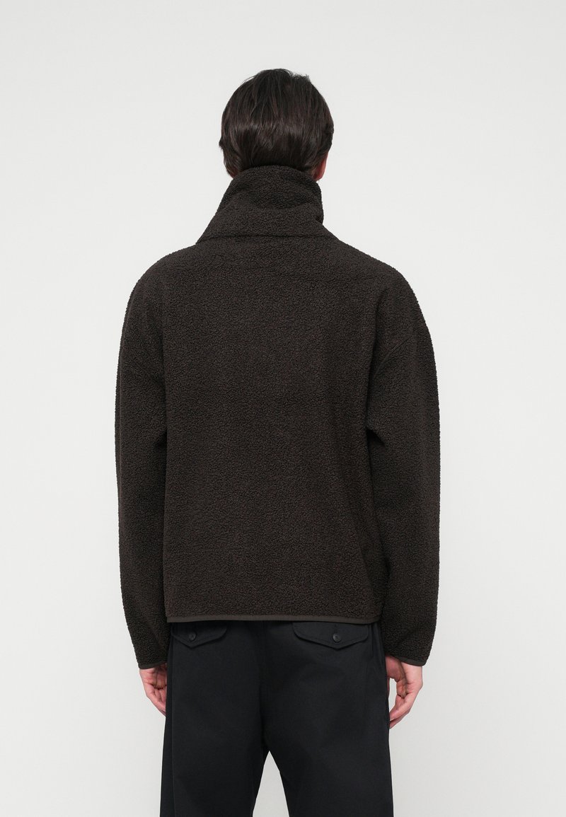 Brun fleecepullover med høj krave, struktureret overflade, dropped shoulders og en lidt løs pasform. Har sorte accenter ved kanten.