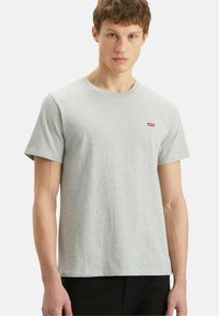 Giovane uomo che indossa una maglietta a maniche corte grigia chiara con un piccolo logo rosso Levi's sul lato sinistro del petto, abbinata a pantaloni neri.