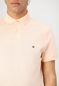 Lyse ferskenfarvet polo-shirt lavet af tekstureret stof, med en klassisk krave, tre knapper og et lille broderet logo på brystet.