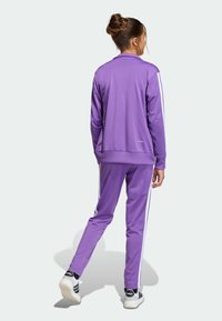 Violetti urheilupuku, joka on tehty sileästä materiaalista ja jossa on valkoiset sivuraitoja. Sisältää vetoketjullisen takin ja kapenevat housut. Kengissä on mustia yksityiskohtia.