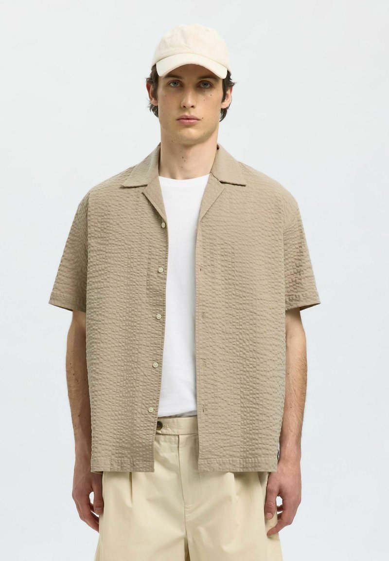 Korte mouwen beiges shirt met een gestructureerd patroon, knoopsluiting en een subtiele kraag, gecombineerd met een wit T-shirt en lichte broek.