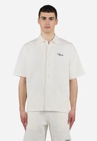 Camicia a maniche corte di colore crema con un motivo a rombi, bottoni frontali e un piccolo logo ricamato in nero.