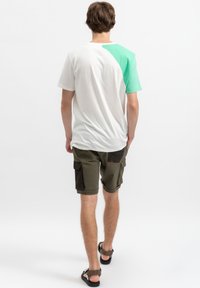 Heren t-shirt met een witte lichaamskleur en een groene linker mouw, gecombineerd met groene cargoshorts met zwarte zakken en opgerolde onderkant, gedragen met sandalen.