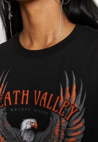 Sort bomulds t-shirt med et grafisk design af en ørn og teksten "Death Valley" i orange og hvid, rund hals, korte ærmer.