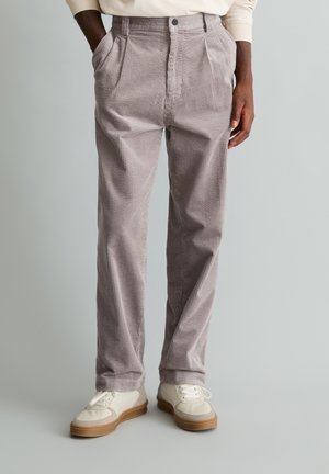 Homme portant un pantalon en velours côtelé gris clair et des baskets blanches, debout les mains dans les poches devant un fond gris uni.