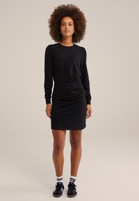 Rochie lungă neagră cu mâneci lungi, talie ajustată și umeri ușor bufanți, dintr-un material elastic. Purtați cu pantofi negri și șosete cu dungi.