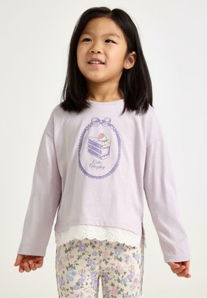 Ung pige med mørkt hår iført en lys lilla langærmet skjorte med kagegrafik og bukser med blomstermønster.