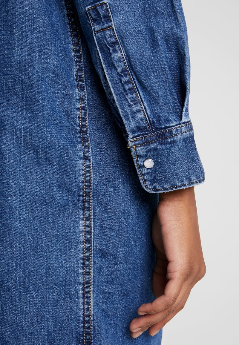 Manche de chemise en denim avec un poignet boutonné. Elle présente un tissu bleu, des coutures visibles et une texture lisse. Une main repose près du bord.