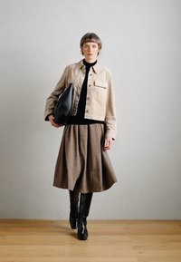 Veste beige cropped sur un col roulé noir, associée à une jupe brune arrivant aux genoux et des bottes noires montantes. Sac noir à la main.