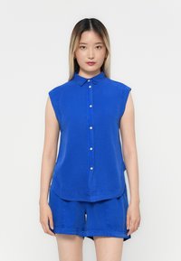 Blaues ärmelloses Hemd mit Knopfleiste, spitzen Kragen und geschwungener Saum. Hergestellt aus weichem Stoff mit subtilen Steppdetails. Kombiniert mit passenden Shorts.