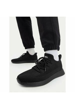 Pies con zapatillas de punto negras con cordones negros y suelas texturizadas, combinadas con calcetines blancos y pantalones jogger negros.