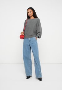 Camisola cinza de mangas compridas com um decote amplo, combinada com jeans de perna larga de um azul claro. Uma pequena mala vermelha acrescenta um toque de cor. Sapatos pretos.