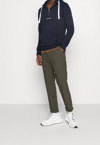 Námořnická mikina na zip s bílými šňůrkami a přední kapsou, doplněná olivově zelenými zúženými kalhotami a bílými teniskami s černými detaily.